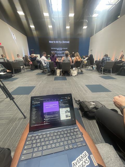 A laptop on someone's lap at the back of a room of people watching a presentation