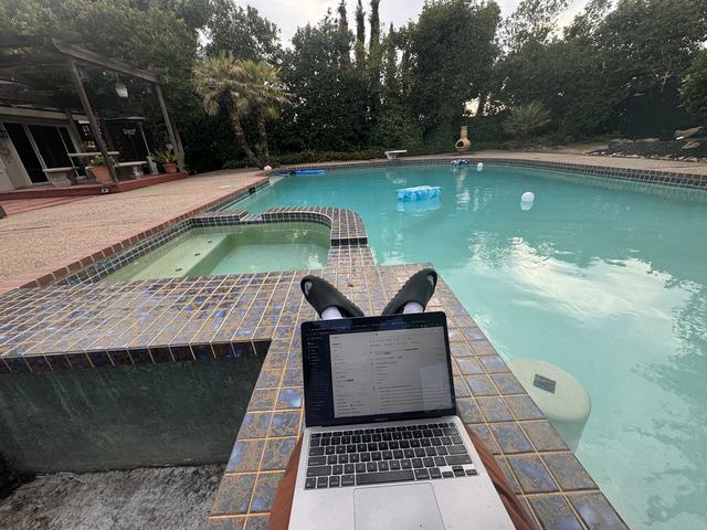 A laptop open on the edge of a backyard pool, feet up beside it
